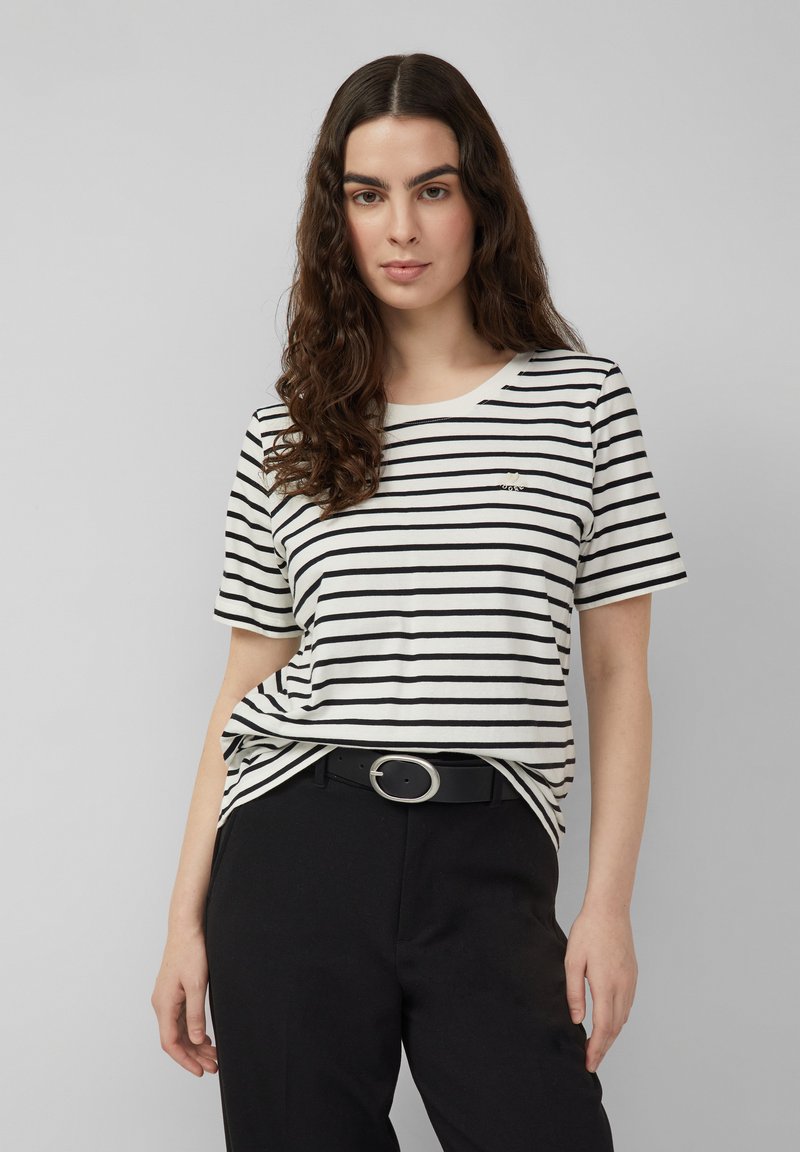 Mujer con cabello largo y rizado de color marrón, lleva una camiseta de manga corta con rayas blancas y negras y pantalones negros con cinturón, de pie frente a un fondo gris.