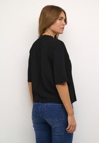 Sort kortærmet bluse med bølget halsudskæring og lodrette striber, parret med mørkeblå jeans, set fra ryggen.