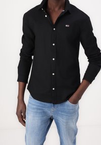 Czarna koszula na guziki z kołnierzykiem, z srebrnymi guzikami i małym logo na piersi. Wykonana z lekkiego materiału, z długimi rękawami.