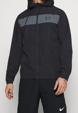Homme portant une veste à capuche zippée noire Under Armour avec une bande grise sur la poitrine et un short noir Nike.