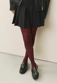 Schwarzer plissierter Rock, bordeauxfarbene Strumpfhosen und schwarze Slipper mit dekorativer Naht. Glattes Leder und matte Oberfläche.