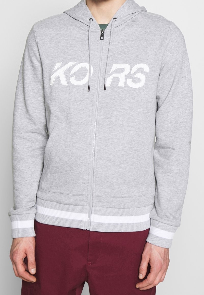 Sweat à capuche gris zippé en tissu doux avec logo blanc "KORS", doté d'une capuche, de poignets côtelés et d'une ceinture à rayures blanches.