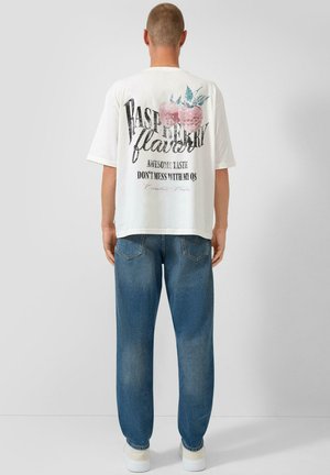 Mann trägt weißes T-Shirt mit pinkem Himbeer-Motiv und Schriftzug, kombiniert mit blauer Jeans und weißen Sneakers, blickt von einer grauen Wand weg.