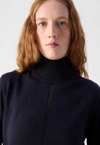Marineblå genser med høy hals laget av myk strikket stoff, med en subtil vertikal sømdetalj foran.