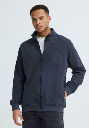 Ung mand med kort krøllet hår iført navy fleecejakke med lynlås og mørke bukser, stående mod en ensfarvet lys baggrund.