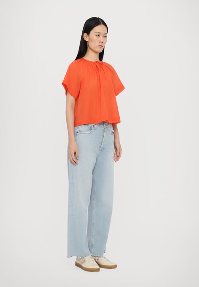 Rotes, kurzes Oberteil mit Rundhalsausschnitt und kurzen Ärmeln. Kombiniert mit hellblauen, weiten Jeans und beigefarbenen Schuhen mit brauner Sohle.