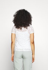 Biała koronkowa bluzka z krótkim rękawem, w kwiatowy wzór, przezroczysty materiał, z falbaniastymi brzegami, nałożona na jasnobrązowy dół.