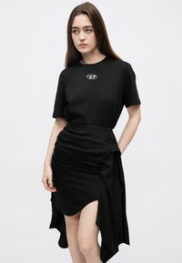 Juoda trumpomis rankovėmis palaidinė su logotipu ant krūtinės, derinama su juoda sijonu, kuriame yra raukiniai ir asimetriškas kraštas.