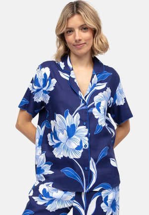 Kobieta nosząca granatową koszulę na krótki rękaw zapinaną na guziki oraz pasujące szorty z dużym białym i niebieskim kwiatowym nadrukiem.