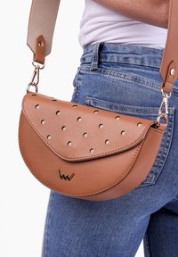 Hnědá kabelka přes rameno z faux kůže s perforovaným vzorem, zlatými detaily a kulatým uzávěrem, nošená na modrých džínech.