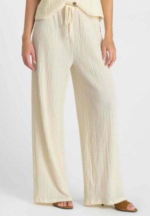 Femme portant un pantalon large texturé crème avec une taille à cordon et des sandales à talon ouvertes marron, debout devant un fond uni.