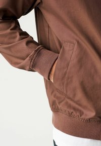 Mano en el bolsillo de una chaqueta marrón con puños y cintura acanalados, llevada sobre una camisa blanca y pantalones oscuros.