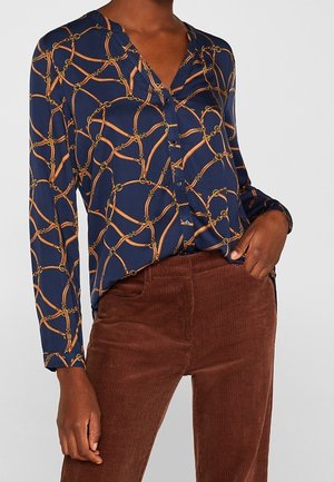 Kobieta w granatowej bluzce z wzorem złotego łańcucha i brązowych spodniach sztruksowych, ręka w kieszeni, na jednolitym tle.