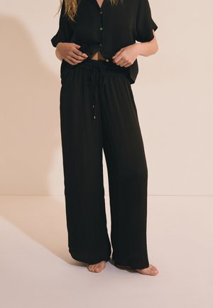 Kobieta w luźnych czarnych spodniach ze sznurkiem oraz krótkim czarnym koszulowym koszulu, stoi boso na jasnej podłodze.