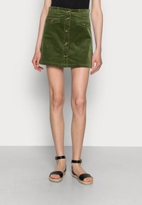 Grön kordflöjelskjorta med framknappar, två fickor och svarta espadrillosandaler med ankelremmar på en neutral bakgrund.