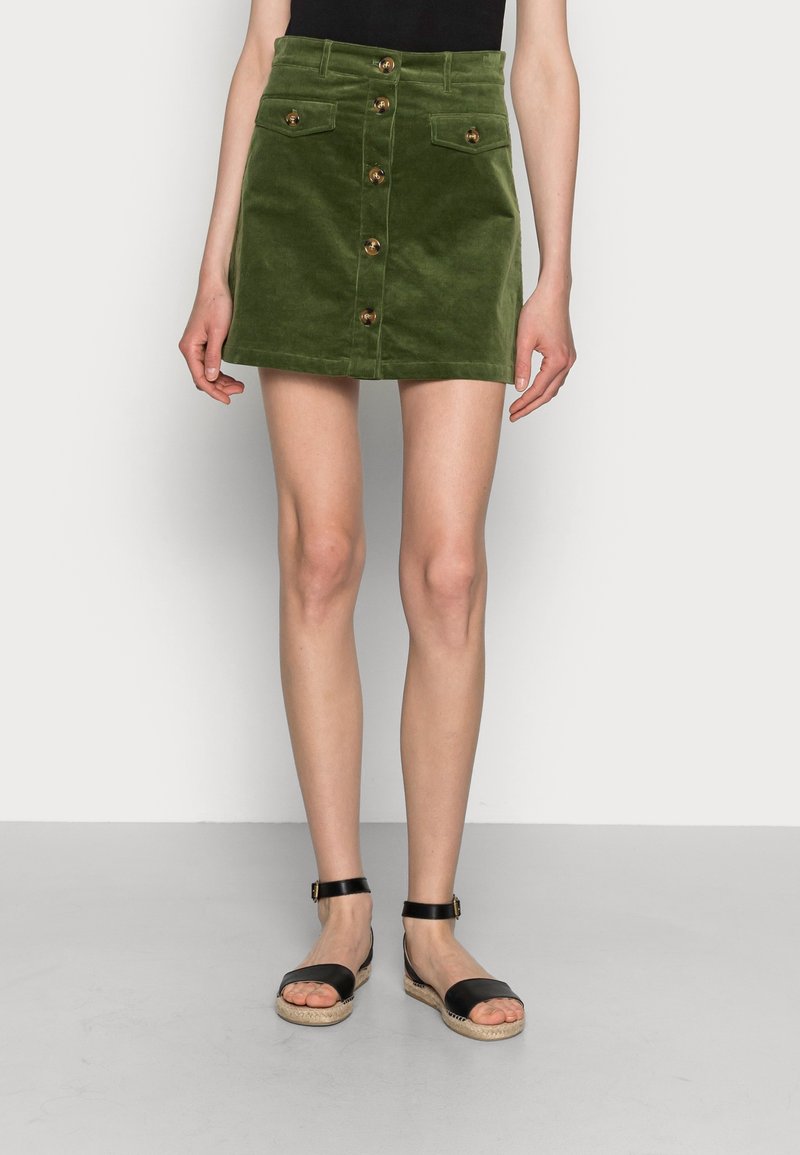 Grön kordflöjelskjorta med framknappar, två fickor och svarta espadrillosandaler med ankelremmar på en neutral bakgrund.
