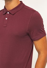 Polo shirt bordeaux realizzato in cotone morbido, caratterizzato da un colletto classico, chiusura con tre bottoni e maniche corte con un design aderente.
