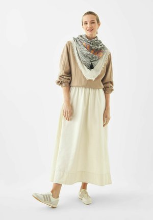 Femme portant un chemisier beige, une longue jupe blanche, une écharpe à motifs avec franges, et des baskets beiges, debout avec un pied légèrement levé.