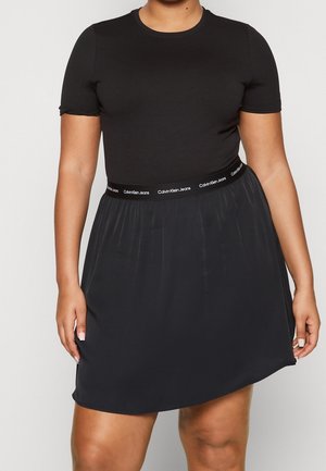 Femme portant un haut noir à manches courtes avec une jupe noire dotée d'une ceinture portant la marque Calvin Klein Jeans.