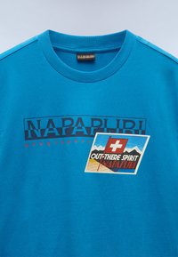 Camiseta de algodón azul con el distintivo texto "NAPAPIJRI" en bold, acentos rojos y un parches gráfico que muestra una bandera suiza y montañas. Diseño de cuello redondo.