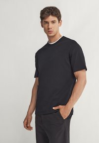 Svart bomullströja med korta ärmar, rundad hals och en vit detalj längs kragen. Modellen poserar på ett lätt sätt med handen i fickan.