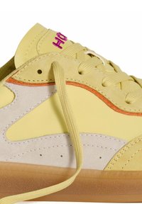 Zapatillas amarillas hechas de materiales de ante y cuero, con un logo rosa, acentos naranjas y una suela texturizada de color marrón claro. Los cordones están desatados.