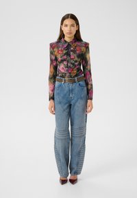 Eine Frau trägt eine farbenfrohe, blumige Bluse mit langen Ärmeln, hochtaillierte blaue Jeans mit gestickten Details, einen braunen Gürtel und burgunderfarbene spitze Schuhe, während sie steht.