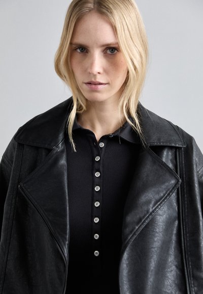 Giacca di pelle nera con ampi rever sopra un polo nero a coste con piccoli dettagli di bottoni. Texture liscia con un design strutturato.