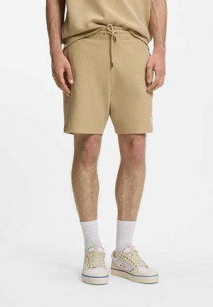 Hombre con pantalones cortos beige con canalé y cordón, calcetines blancos y zapatillas crema sobre fondo blanco liso.