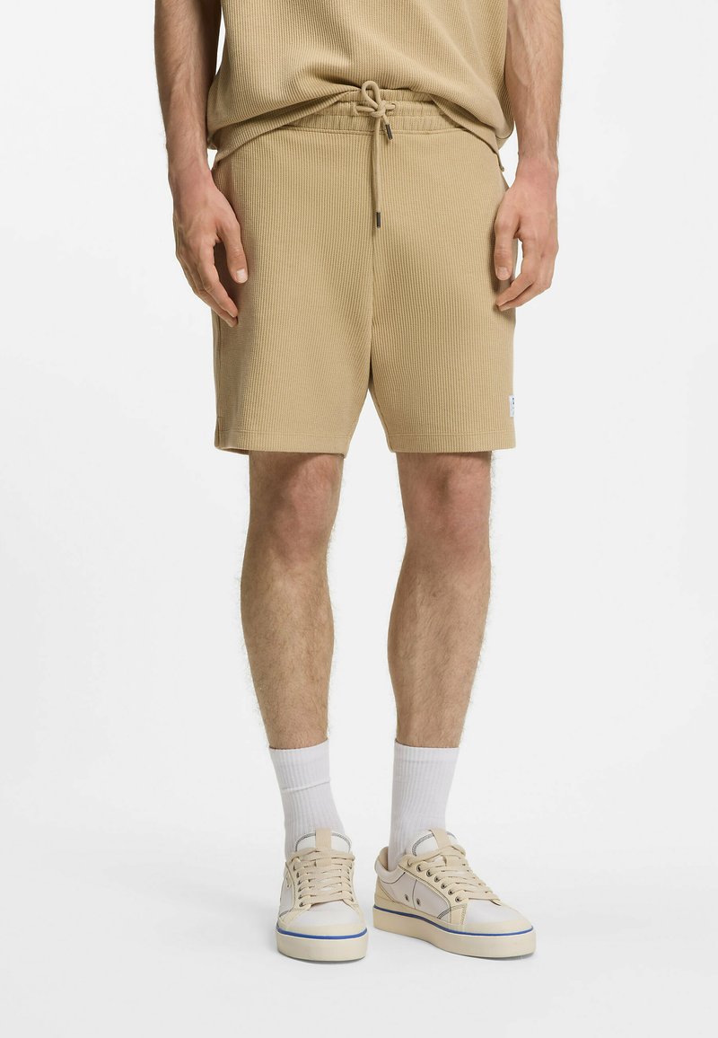 Homme portant un short beige côtelé avec un cordon de serrage, des chaussettes blanches et des baskets crème sur fond blanc uni.