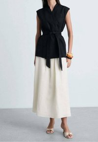 Top senza maniche in lino nero con design avvolgente e vita legata, abbinato a una maxi gonna bianca fluida e sandali beige. Texture morbide.