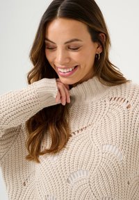 Pull en maille beige clair avec col haut, présentant des motifs texturés en vagues et des découpes à l'avant. Matériau doux et épais.