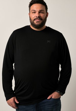 Schwarzes Langarmshirt aus glattem Stoff, mit dezentem Logo auf der Brust und lässigem Schnitt. Kombiniert mit dunkler Denim-Jeans.