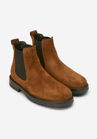 Bottines Chelsea en daim marron avec des panneaux élastiques noirs sur les côtés, orteils ronds et semelles texturées. Dotées de languettes à l'arrière pour un enfilage facile.