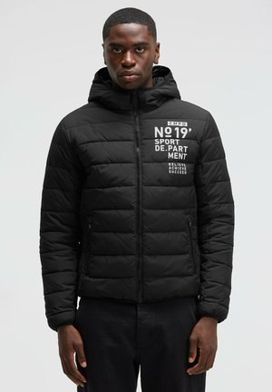 Young man wearing a black padded hooded jacket with white sports text on the chest, standing against a plain light background.