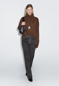 Maglione lavorato a maglia marrone, pantaloni in denim grigio scuro, borsa a spalla nera, collana con pendente in argento, modello che cammina su uno sfondo bianco semplice.