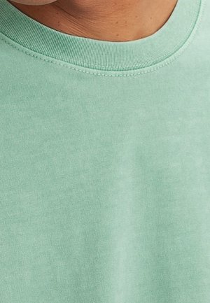 Gros plan d'une personne portant un t-shirt en coton vert clair à col rond montrant la texture du tissu et les coutures de l'encolure.