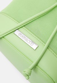 Dettaglio di una borsa a tracolla verde chiaro con tessuto a rete, rifiniture simili alla pelle e placca del marchio "Even&Odd" in argento con tracolla annodata.
