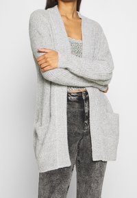 Lichtgrijze gebreide cardigan met een open voorkant, geribbelde textuur en twee voorzakken. G gedragen over een gemusterde top en donkere jeans.