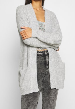 Cardigan grigio chiaro in maglia con apertura frontale, tessuto a coste e due tasche anteriori. Indossato sopra una maglietta a fantasia e jeans scuri.