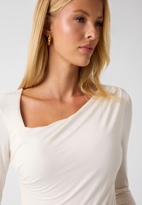 Cremefarbener Langarmtop mit drapiertem Ausschnitt, weicher Textur und figurbetonter Silhouette. Mit einem quadratisch geschnittenen Design an einer Schulter.