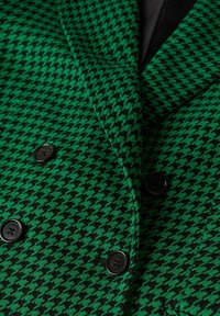 Blazer à motif pied-de-poule vert et noir en tissu texturé, avec des boutons noirs et une coupe croisée.