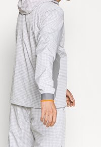 Veste à capuche blanche à motif quadrillé avec des accents gris, dotée de poignets élastiques et d'une bordure orange, conçue pour une coupe décontractée.