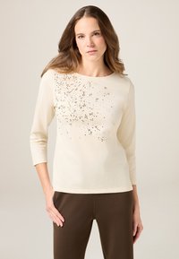 Crème lange mouwen top met een ronde halslijn, voorzien van een verspreid goudkleurig pailletontwerp aan de voorkant; gecombineerd met een bruine broek.