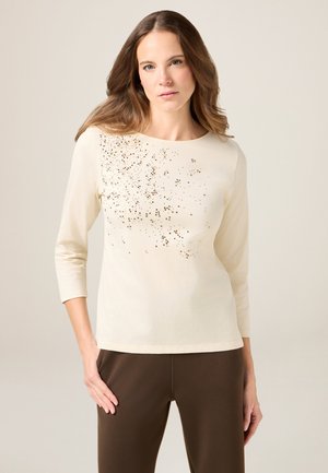 Kremowa bluzka z długim rękawem, okrągłym dekoltem i rozsypanym złotym cekinowym wzorem z przodu; zestawiona z brązowymi spodniami.