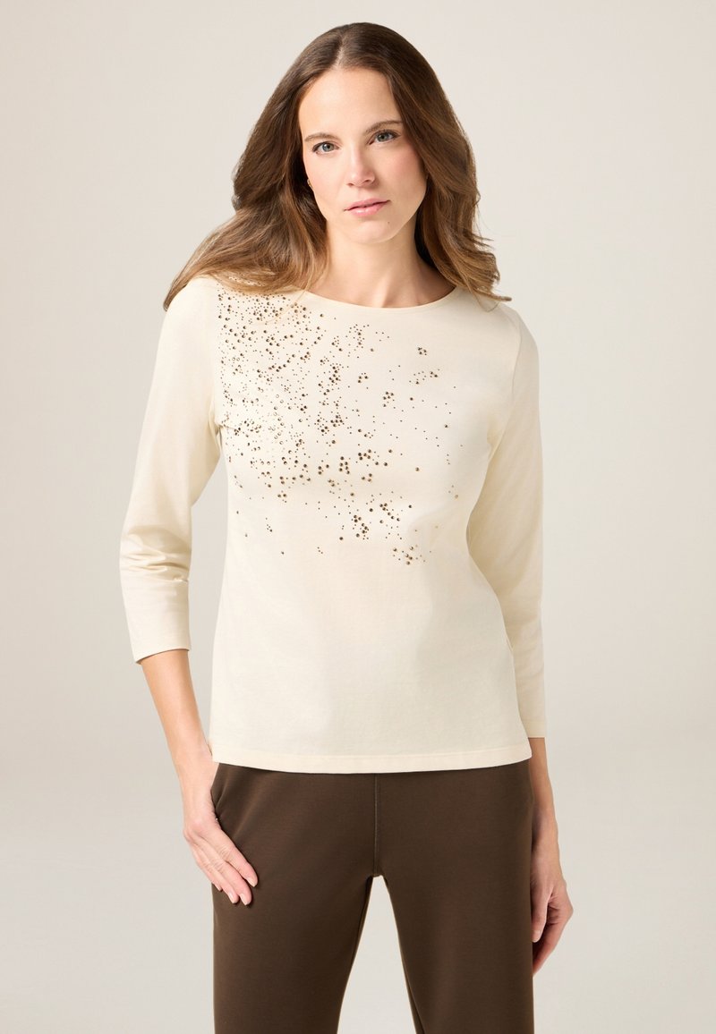 Crème lange mouwen top met een ronde halslijn, voorzien van een verspreid goudkleurig pailletontwerp aan de voorkant; gecombineerd met een bruine broek.