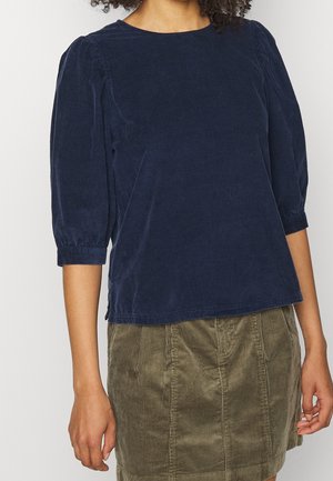 Vrouw die een marineblauwe velvet top met lange mouwen en een olijfgroene ribfluwelen rok draagt, afgebeeld van schouders tot halverwege de dijen.
