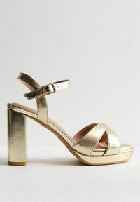 Sandalias de tacón alto metálicas doradas con tira al tobillo, punta abierta y diseño entrecruzado. Tacón ancho con suela texturizada.