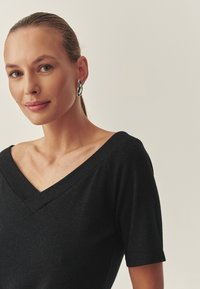 Čierny top s výstrihom do V a krátkymi rukávmi, s jemným leskom a hladkou textúrou. Výrazné strieborné náušnice dopĺňajú vzhľad.