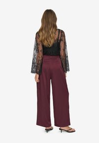 Woman with long wavy hair wearing black lace sheer top, high-waisted wide-leg burgundy pants, and black heeled sandals facing away.